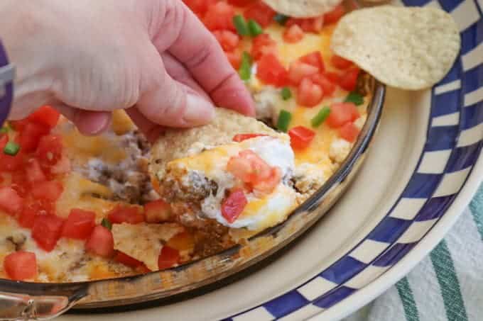 This Taco Dip is an easy game day dish to make, and fun to serve. The four layers of deliciousness are sure to please your hungry crowd. This dip of refried beans, taco seasoned ground beef, sour cream and Mexican cheese topped with diced tomato, jalapeño, and served with tortilla chips is a fan favorite at our house!