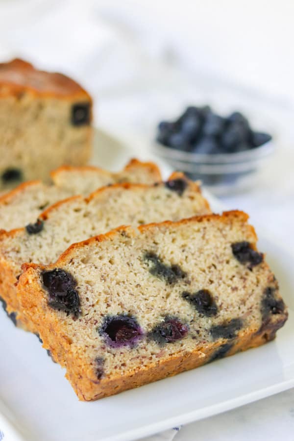 Slices of quick bread, blueberry banana bread on a plate with blueberries behind it.