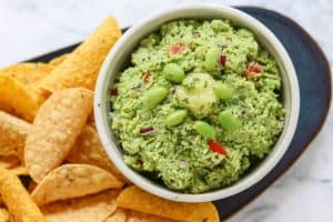 A bowl of guacamole with edamame and ginger surrounded by chips.