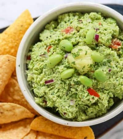 A bowl of guacamole with edamame and ginger surrounded by chips.