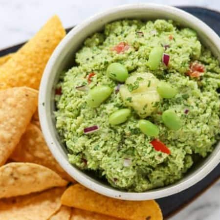 A bowl of guacamole with edamame and ginger surrounded by chips.