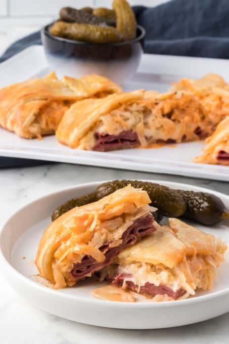 Pickles and a puff pastry corned beef sandwich on a plate in the foreground with more slices behind it.