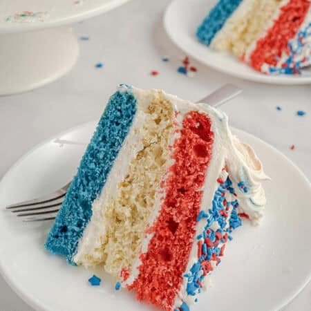 A cake slice with a blue, white, and red layer on a plate with part of the American flag in the foreground.