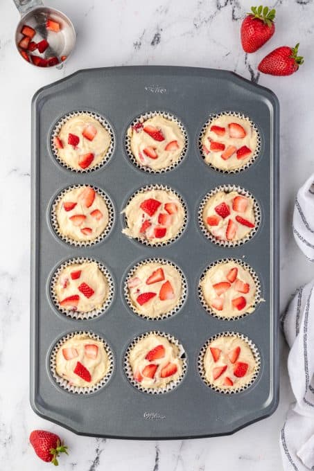 A muffin tin filled with batter and fresh strawberries.