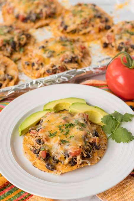 A tostada topped with chicken and cheese served with slices of avocado.