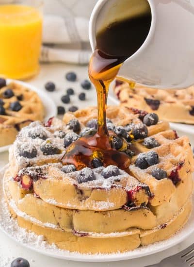 Syrup being poured on a stack of waffles made with blueberries.