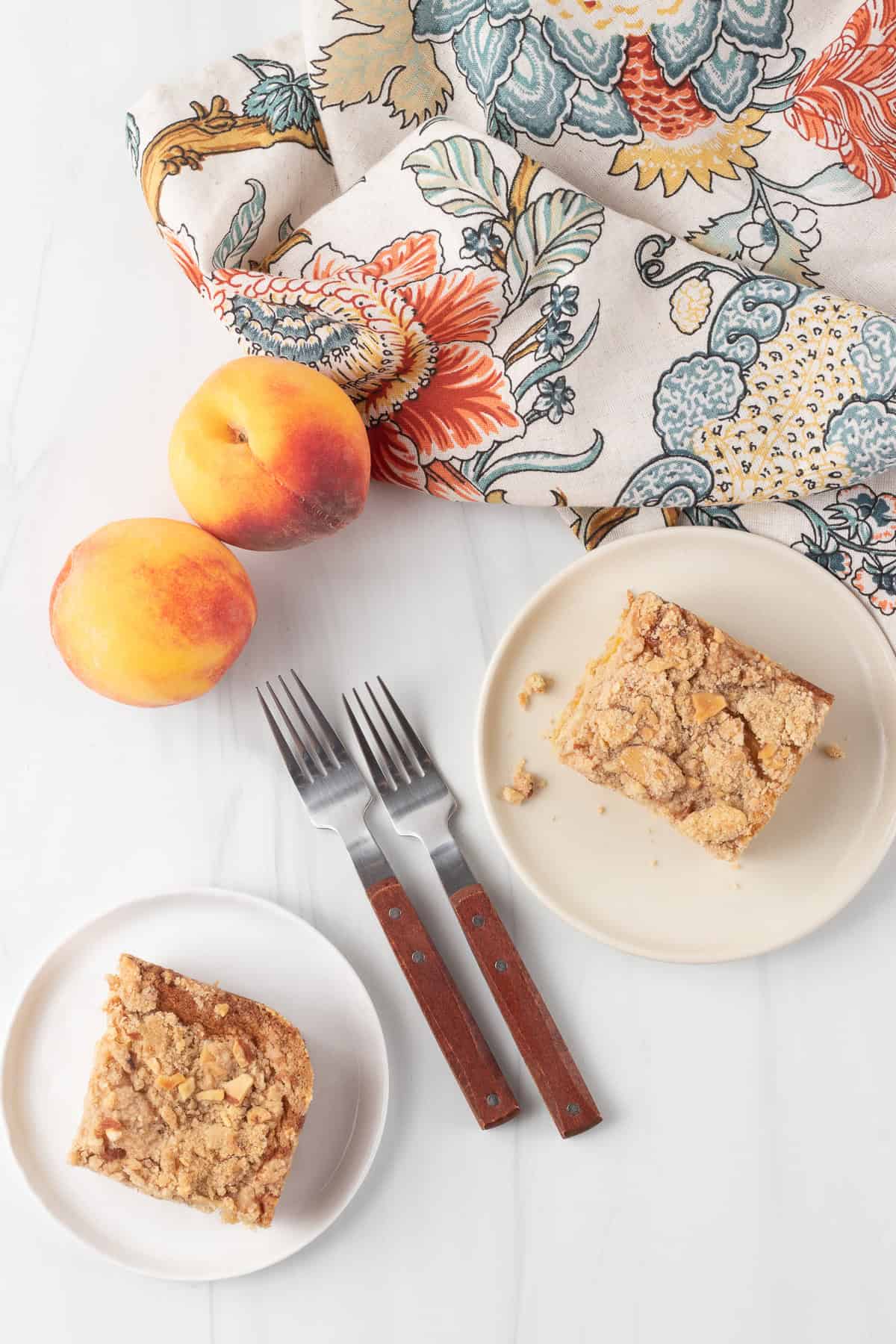 Looking down at two slices of cake on separate plates with two forks, two peaches and a floral napkin.