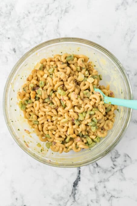 Pasta, celery, chopped hard-boiled eggs, red onion, and green pepper mixed together in a glass bowl with a light green spatula.