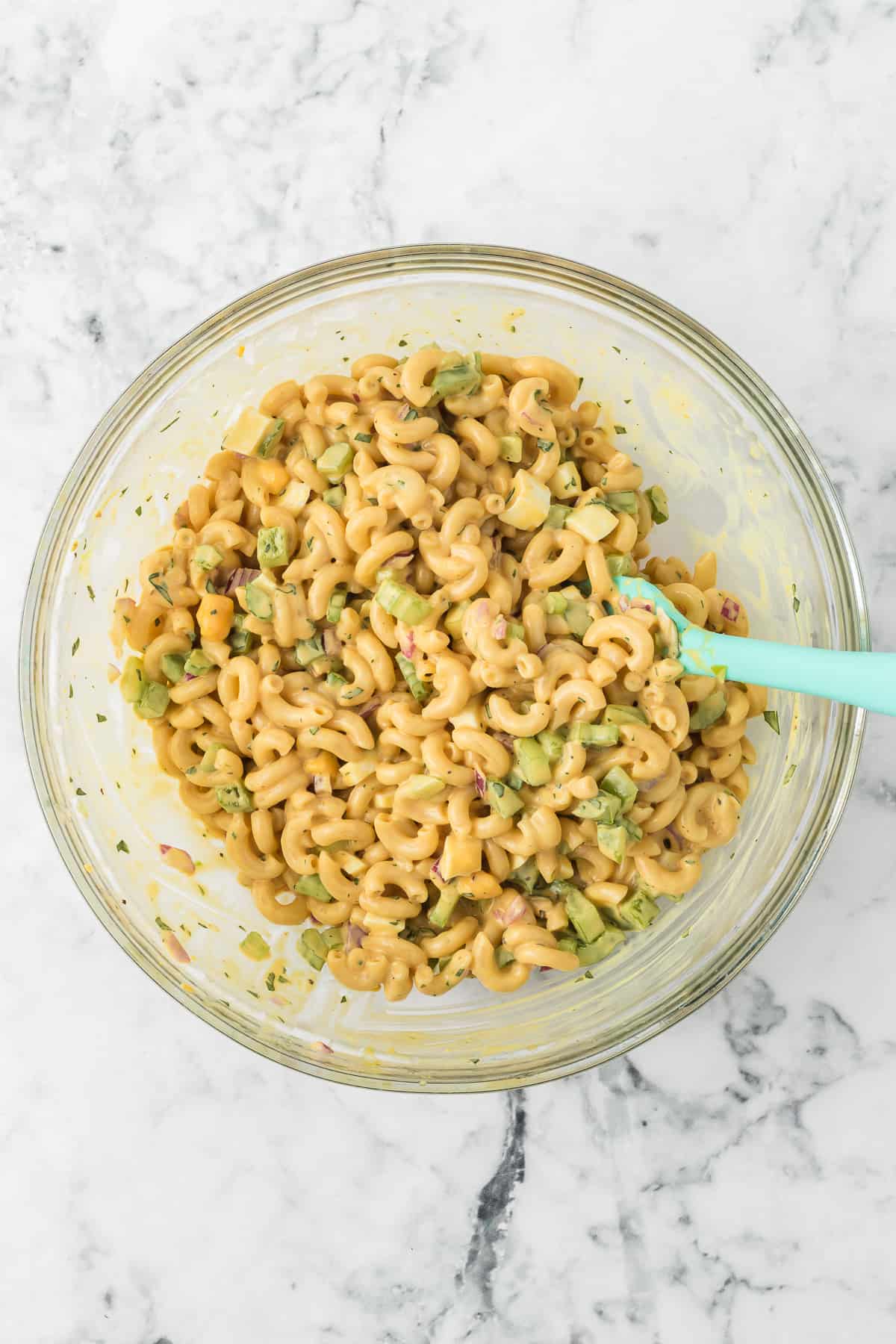 Pasta, celery, chopped hard-boiled eggs, red onion, and green pepper mixed together in a glass bowl with a light green spatula.